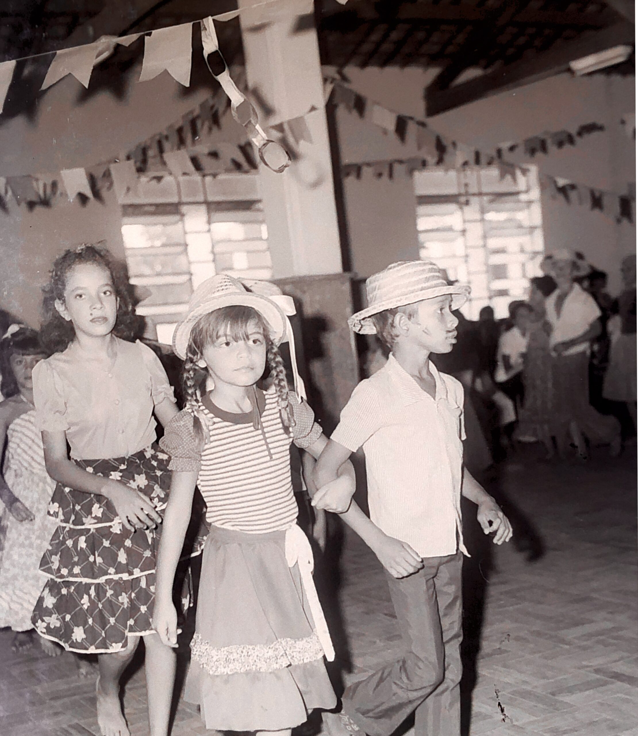 Fotografia em preto e branco de uma quadrilha junina infantil realizada em ambiente fechado na cidade de Barbalha. Três crianças aparecem em destaque, caminhando lado a lado em fila. Um menino, usando camisa clara e chapéu de palha, conduz uma menina vestida com roupa típica de festa junina, incluindo saia rodada, blusa listrada e chapéu com tranças postas sobre os ombros. Atrás deles, outra menina com vestido estampado segue descalça. O espaço está decorado com bandeirolas penduradas no teto e correntes de papel colorido, típicas de festas juninas. Ao fundo, outros participantes e observadores assistem à dança. Registro feito na década de 1980 pelo Padre Paulo, na cidade de Barbalha.