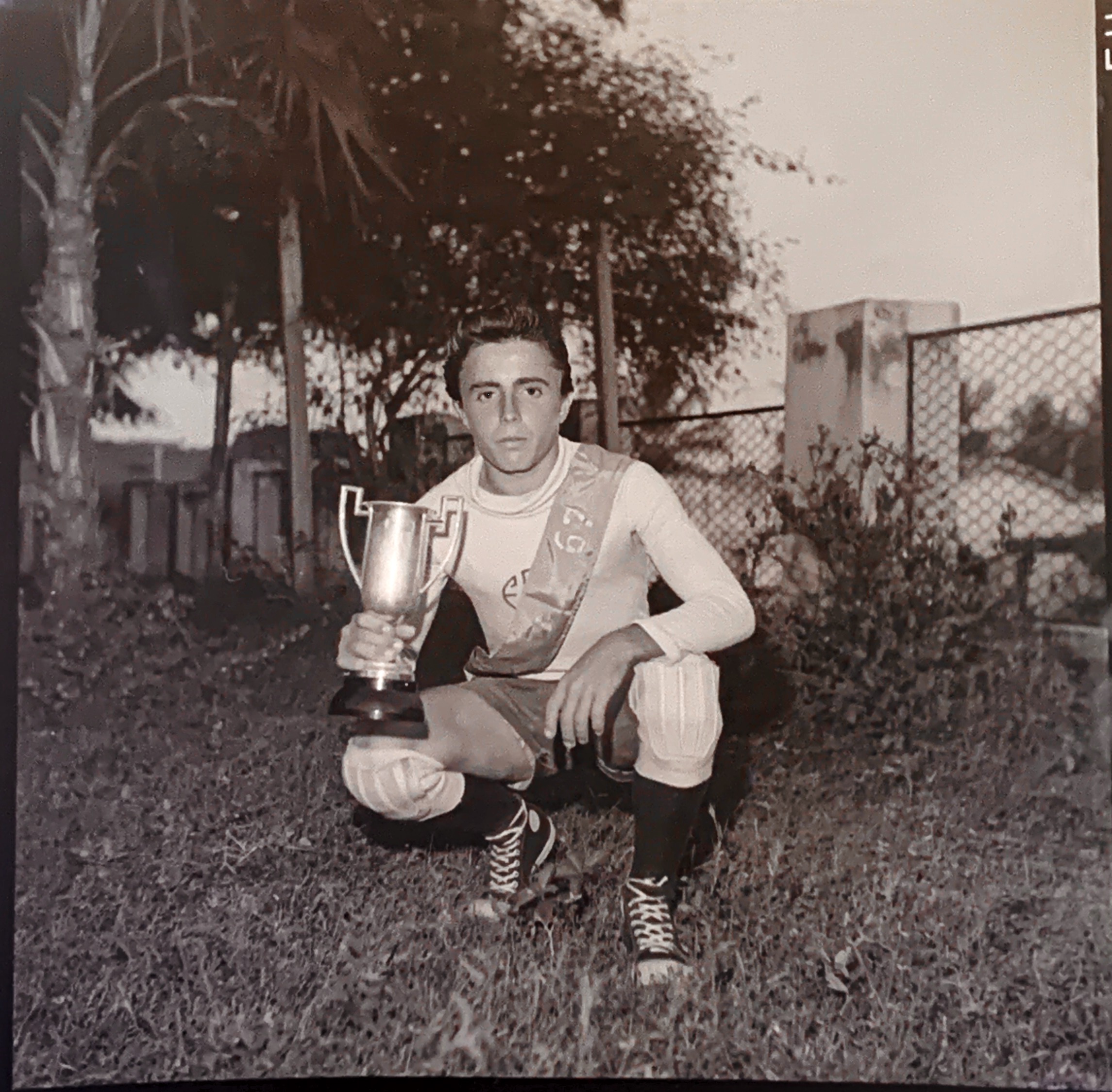 Jovem agachado sobre a grama segura um troféu com as duas mãos. Ele veste uniforme esportivo com a sigla “ESC” estampada na camisa e uma faixa diagonal com os dizeres “REPÚBLICA” e “1967”. Usa joelheiras, meias altas e tênis de cano longo, indicando participação em atividade esportiva. Ao fundo, há vegetação, palmeiras e uma cerca de arame com edificações residenciais visíveis. Fotografia registrada em Barbalha por Padre Paulo.