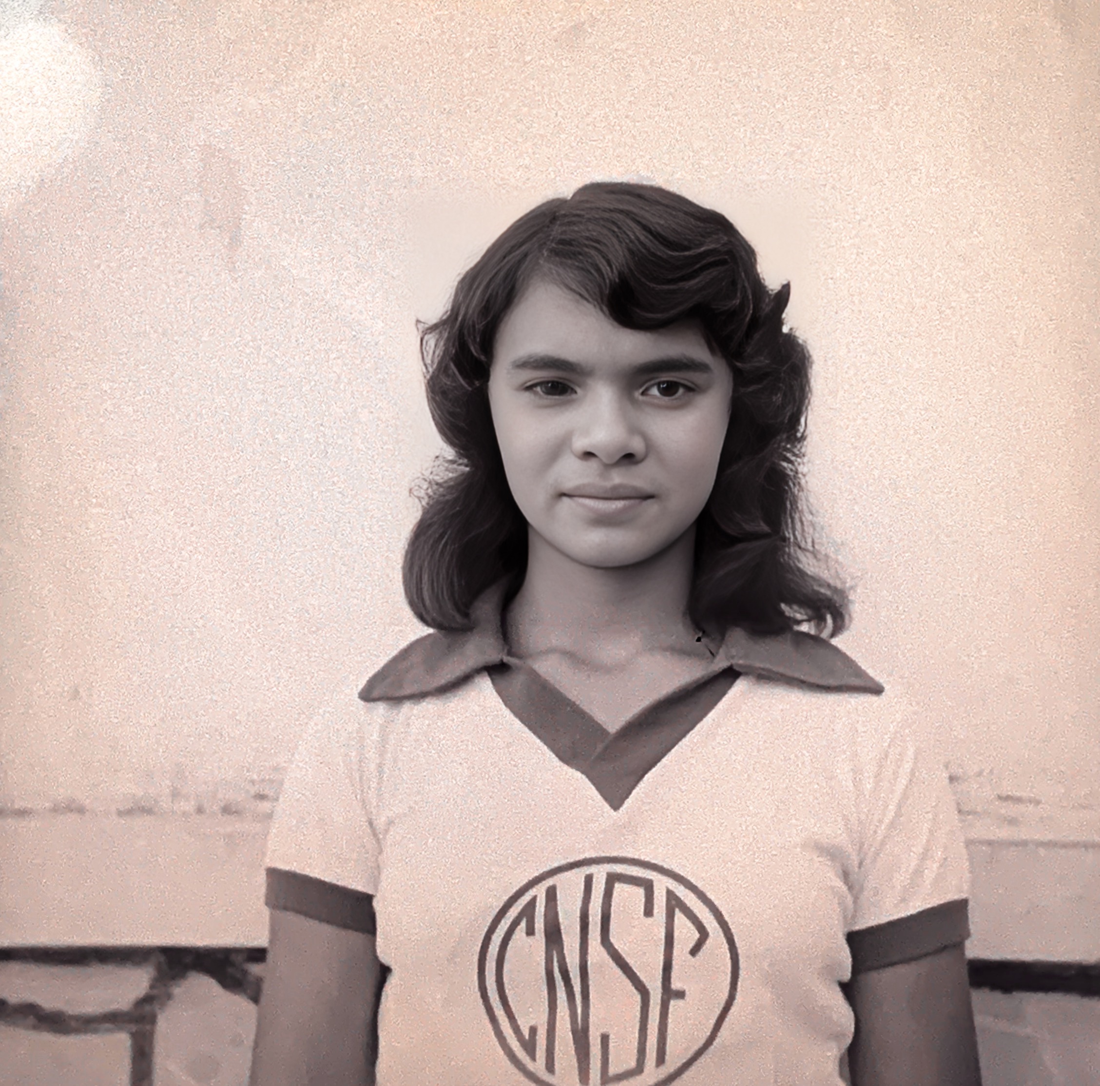 Menina em Barbalha, posando diante de uma parede clara. Ela veste uniforme escolar com o emblema “CNSF” estampado na camisa. Fotografia de Padre Paulo.